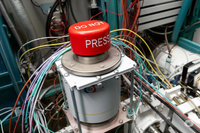 A big red button labelled DO NOT PRESS is attached to the top of an ominous looking device to which many wires and conduits are attached.